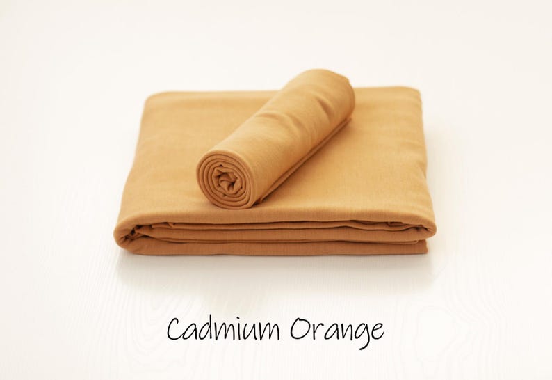 May include: A cadmium orange fabric cloth folded into a rectangular shape with a smaller matching piece rolled and placed on top. The text Cadmium Orange is printed in a simple black font below the fabric, indicating the specific color name.