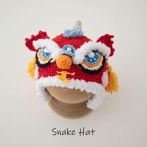 May include: Red, white and yellow crocheted lion dance hat with a white fur trim. The hat has embroidered eyes and a nose. The hat is on a wooden display stand. Snake Hat.