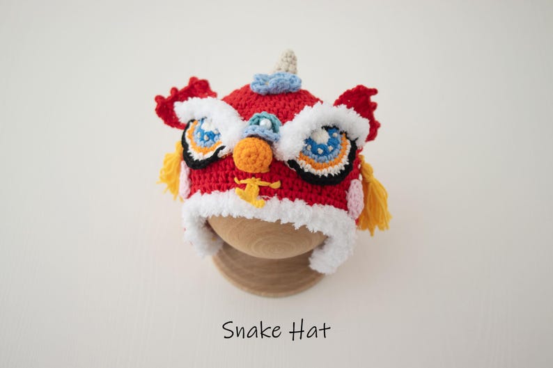 May include: Red, white and yellow crocheted lion dance hat with a white fur trim. The hat has embroidered eyes and a nose. The hat is on a wooden display stand. Snake Hat.