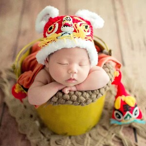 May include: A newborn baby wearing a red and white lion dance hat is sleeping in a yellow bucket. The baby is surrounded by a brown and beige fabric.