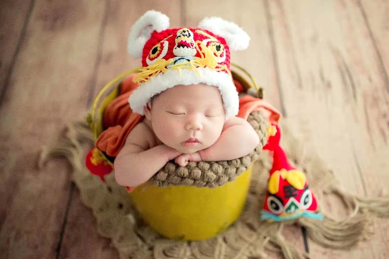 May include: A newborn baby wearing a red and white lion dance hat is sleeping in a yellow bucket. The baby is surrounded by a brown and beige fabric.