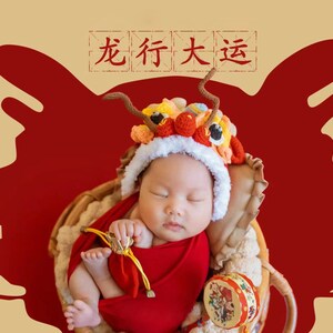 May include: A baby wearing a red and yellow lion costume sleeps in a wicker basket against a red background. The text "龙行大运" is written in black on a white background above the baby.