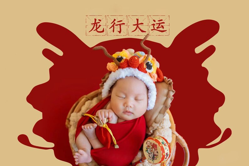 May include: A baby wearing a red and yellow lion costume sleeps in a wicker basket against a red background. The text "龙行大运" is written in black on a white background above the baby.