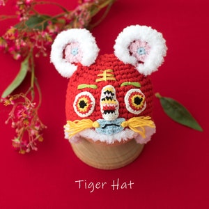 May include: Red crocheted tiger hat with white fur trim, yellow yarn whiskers, and embroidered eyes. The hat is on a wooden stand against a red background. "Tiger Hat" is written below the hat.