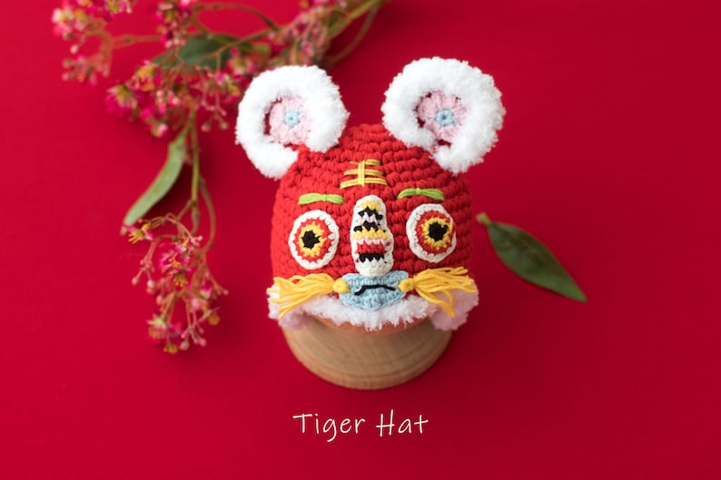 May include: Red crocheted tiger hat with white fur trim, yellow yarn whiskers, and embroidered eyes. The hat is on a wooden stand against a red background. "Tiger Hat" is written below the hat.