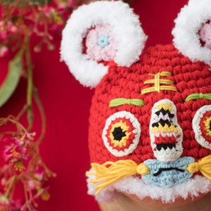 May include: Red crocheted lion hat with white fur trim, yellow yarn tassels, and embroidered details. The hat features a lion face with black eyes, a red nose, and a white mouth.