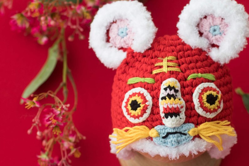 May include: Red crocheted lion hat with white fur trim, yellow yarn tassels, and embroidered details. The hat features a lion face with black eyes, a red nose, and a white mouth.