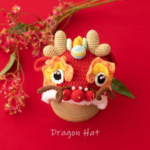 May include: A red, yellow and brown crocheted dragon hat with white fur trim. The hat has large black eyes and a small, blue and white ball on top. The hat is sitting on a wooden base against a red background.