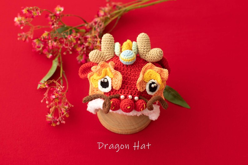 May include: A red, yellow and brown crocheted dragon hat with white fur trim. The hat has large black eyes and a small, blue and white ball on top. The hat is sitting on a wooden base against a red background.