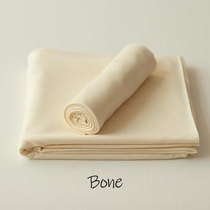 May include: A folded piece of solid bone-colored fabric with a smooth texture, featuring a rolled section resting on top of the flat base. The word Bone is printed in black text below the fabric, indicating the specific color of the material.