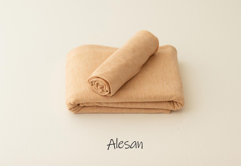May include: A folded piece of light tan fabric with a smooth, soft texture. A smaller, rolled section of the same tan material rests on top of the larger folded base. The word Alesan is printed in black text below the fabric pieces.