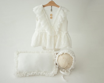 May include: A white lace baby outfit with a ruffled neckline and a matching hat and pillow. The outfit has two button closures at the neckline.