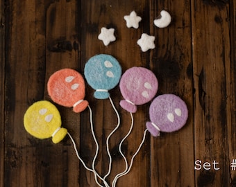 May include: Set of five felt balloons in yellow, orange, blue, purple and pink colors with white felt dots. The balloons are attached to white strings and are arranged in a semi-circle on a brown wooden background. There are also five white felt stars and a white felt crescent moon on the background. The text "Set #1" is written in white on the bottom right corner of the image.