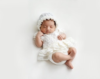 May include: A sleeping baby wearing a white lace dress with ruffled layers and a matching bonnet. The bonnet and dress have delicate lace details. The baby is lying on a white surface.