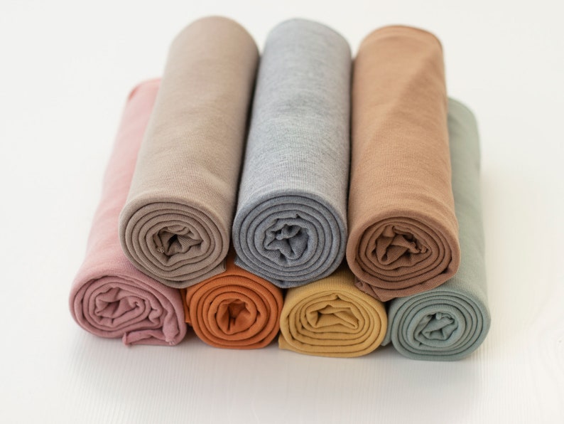 May include: A collection of seven fabric rolls arranged in a stack, featuring a variety of solid colors including dusty pink, beige, light gray, tan, terracotta, mustard yellow, and sage green. Each piece of fabric is tightly rolled into a cylinder.