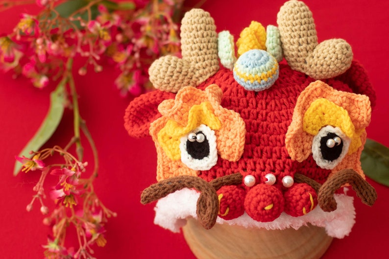 May include: A red, orange and yellow crocheted lion dance hat with white fur trim and pearl-like beads. The hat has a small, multi-coloured ball on top.