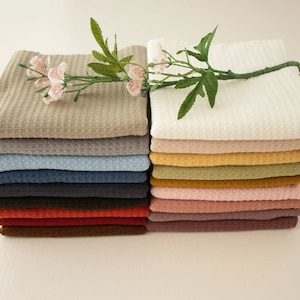 Könnte beinhalten: Stapel von Waffelstoff in verschiedenen Farben, darunter Beige, Blau und Rotbraun. Ein Zweig mit kleinen rosa Blüten liegt auf dem Stoff. Der Stoff hat ein strukturiertes, gitterartiges Muster.
