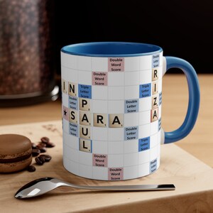 May include: A white ceramic mug with a blue rim and handle. The mug features a Scrabble board design with the word "PAUL" spelled out in the center. The letters are on tiles with the corresponding point values.