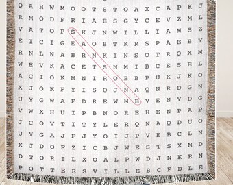 May include: A white woven throw blanket with a word search puzzle featuring the word "Drew Me" highlighted in pink. The puzzle includes letters in a grid pattern.