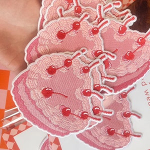 May include: A stack of pink and white embroidered iron-on patches featuring a pink cake with red cherries. The text on the card reads "Have your cake and wear it too."