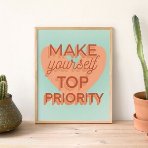 May include: A framed print with a light blue background and a peach-colored heart shape with the text "MAKE yourself TOP PRIORITY" in a layered, 3D-style font.