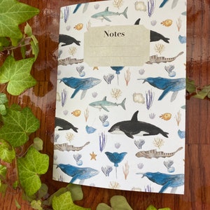 May include: A white notebook with a colorful ocean-themed design. The cover features whales, sharks, fish, and sea plants. The notebook has a label that says "Notes".