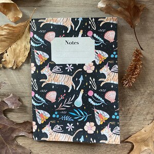 May include: A black notebook with a whimsical design featuring tigers, butterflies, flowers, and stars. The cover has the word "Notes" printed on it.