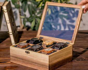 May include: Wooden watch display case with a glass top, holding six watches with various wood tones and band colors. The case is open, revealing the watches. Two leather-bound books are in the background.