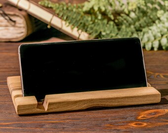 May include: A wooden phone stand with a black smartphone resting on it. The stand is made of light brown wood and has a simple design. The stand is sitting on a dark brown wooden surface.
