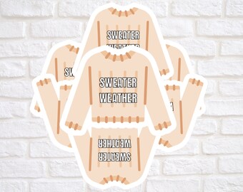 May include: A collection of beige sweater-shaped stickers. Each sticker has the words "SWEATER WEATHER" in black block letters. The stickers are arranged in a cluster against a white brick wall background.