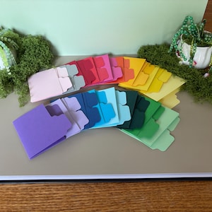 May include: A collection of multi-colored cardstock file folders arranged in a fan shape. The set includes various shades of pink, red, orange, yellow, purple, blue, and green, each featuring a single tab on the top edge for organization.