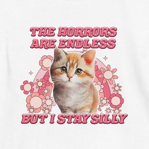 Puede incluir: Camiseta blanca con un gato de dibujos animados en naranja y blanco, sobre un diseño floral y arcoíris rosa. El texto "THE HORRORS ARE ENDLESS BUT I STAY SILLY" está impreso encima y debajo del gato.