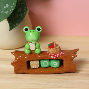 May include: A whimsical ceramic perpetual calendar featuring a green frog holding a sandwich, a picnic basket with a red apple, and date blocks. The calendar displays "JAN 09" on a brown log-shaped base. The frog has large black eyes.