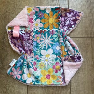 Puede incluir: Una manta de seguridad cuadrada para bebé con un diseño de patchwork de varios patrones florales en turquesa, amarillo, morado y rosa. La manta tiene un reverso de felpa rosa claro texturizado, una cinta de satén rosa y una etiqueta de tela.