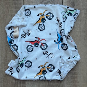 Op de afbeelding: Een witte babydeken met een zachte, pluche rand. De deken heeft kleurrijke illustraties van crossmotoren in verschillende kleuren, geblokte vlaggen en zwarte sterren. Een satijnen lint is aan de deken bevestigd.