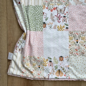May include: A patchwork blanket featuring a variety of square panels with patterns of butterflies, beetles, bees, and floral sprigs in soft shades of pink, sage green, and yellow. The fabric includes polka dots, chevron stripes, and a white tag labeled Rosie B.