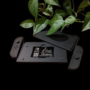Puede incluir: Accesorios negros para consola Nintendo Switch con temática navideña. El conjunto incluye un soporte para consola, dos mandos Joy-Con y un tarjetero de juego. El tarjetero de juego tiene el texto "Merry Christmas". Hojas verdes en el fondo.