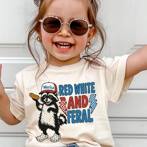 May include: A young child wearing a cream-colored t-shirt with a graphic of a raccoon holding a hot dog and the words "Red White and Feral." The child is wearing sunglasses and making a peace sign with both hands.