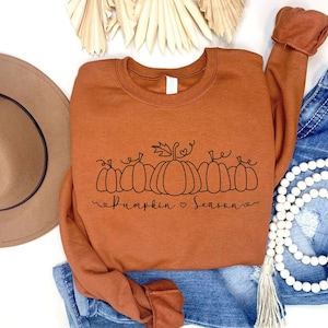 May include: A brown sweatshirt with a black graphic of pumpkins and the text "pumpkin season". The sweatshirt is laid out on a white surface with a brown hat, blue jeans, and a white beaded necklace.