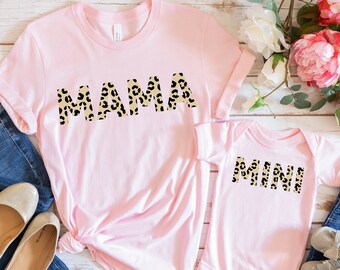 May include: Pink t-shirts with the words "MAMA" and "MINI" in leopard print. The shirts are on a white wooden background with jeans and shoes.