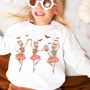 May include: White sweatshirt featuring a Halloween-themed design of three skeleton ballerinas in pink tutus, holding jack-o'-lantern buckets, and dancing under bats. The design is on a white sweatshirt.