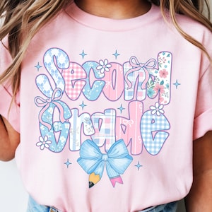 May include: Pink short-sleeved t-shirt with the words "Second Grade" in a patchwork design. The letters are decorated with floral patterns, bows, and a blue pencil. The shirt is worn by a child.