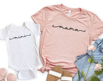 May include: A pink t-shirt with the word "mama" written in black cursive script, and a white onesie with the word "mini" written in black cursive script. The onesie is paired with pink baby shoes. The t-shirt is paired with blue jeans and brown sandals.