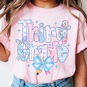 May include: A light pink t-shirt featuring the text "Third Grade" in a large, multi-patterned font with blue and pink plaid, gingham, and floral designs. The graphic includes decorative bows, small flowers, sparkles, and a pencil icon.