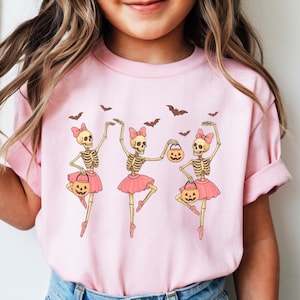May include: Pink t-shirt featuring three dancing skeleton ballerinas with pink tutus and bows, holding jack-o'-lantern treat bags. Bats are flying above them. A fun Halloween-themed graphic tee for kids.