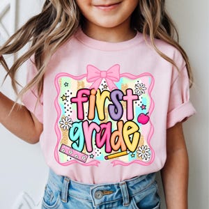 May include: Pink short-sleeved t-shirt with the words "first grade" in colorful letters. The design includes a pink bow, daisies, stars, a ruler, a pencil, and a pink apple. The shirt is suitable for a child.