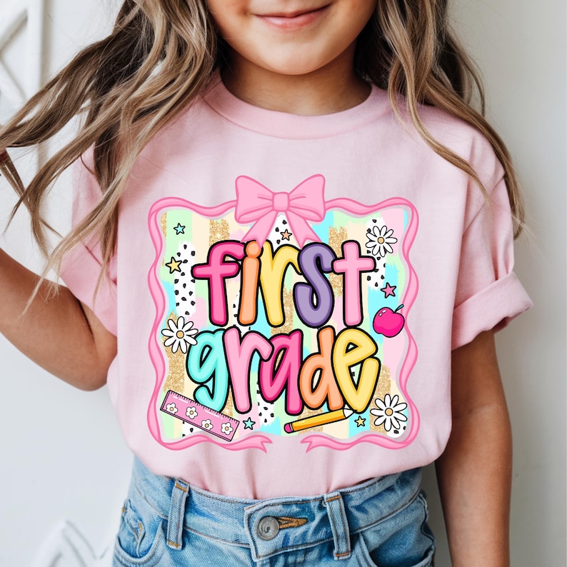 May include: Pink short-sleeved t-shirt with the words "first grade" in colorful letters. The design includes a pink bow, daisies, stars, a ruler, a pencil, and a pink apple. The shirt is suitable for a child.