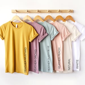 May include: A row of eight t-shirts hanging on a wooden rack. The t-shirts are in various colors, including yellow, pink, blue, peach, beige, and white. The colors of the t-shirts are labeled with their names: Heather Mustard, Heather Orchid, Heather Dusty Blue, Heather Peach, Natural, White.