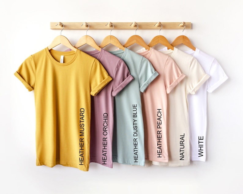 May include: A row of eight t-shirts hanging on a wooden rack. The t-shirts are in various colors, including yellow, pink, blue, peach, beige, and white. The colors of the t-shirts are labeled with their names: Heather Mustard, Heather Orchid, Heather Dusty Blue, Heather Peach, Natural, White.
