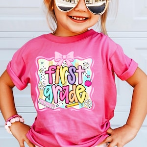 May include: A pink t-shirt with the words "First Grade" in colorful bubble letters. The graphic includes a pink bow, stars, flowers, a pencil, and an apple. The shirt is worn by a child wearing sunglasses and a pink bracelet.